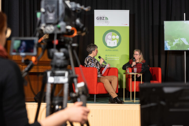 Ein Presse-Foto, auf dem auf einer Bühne eine Botschafterin für Alphabetisierung und Teilhabe von einer Moderatorin interviewt wird.