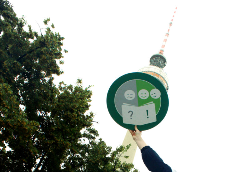 Ein Presse-Foto mit einem runden Siegel, dem Alpha-Siegel, das von einer Hand vor die Kuppel des Berliner Fernsehturms gehalten wird.